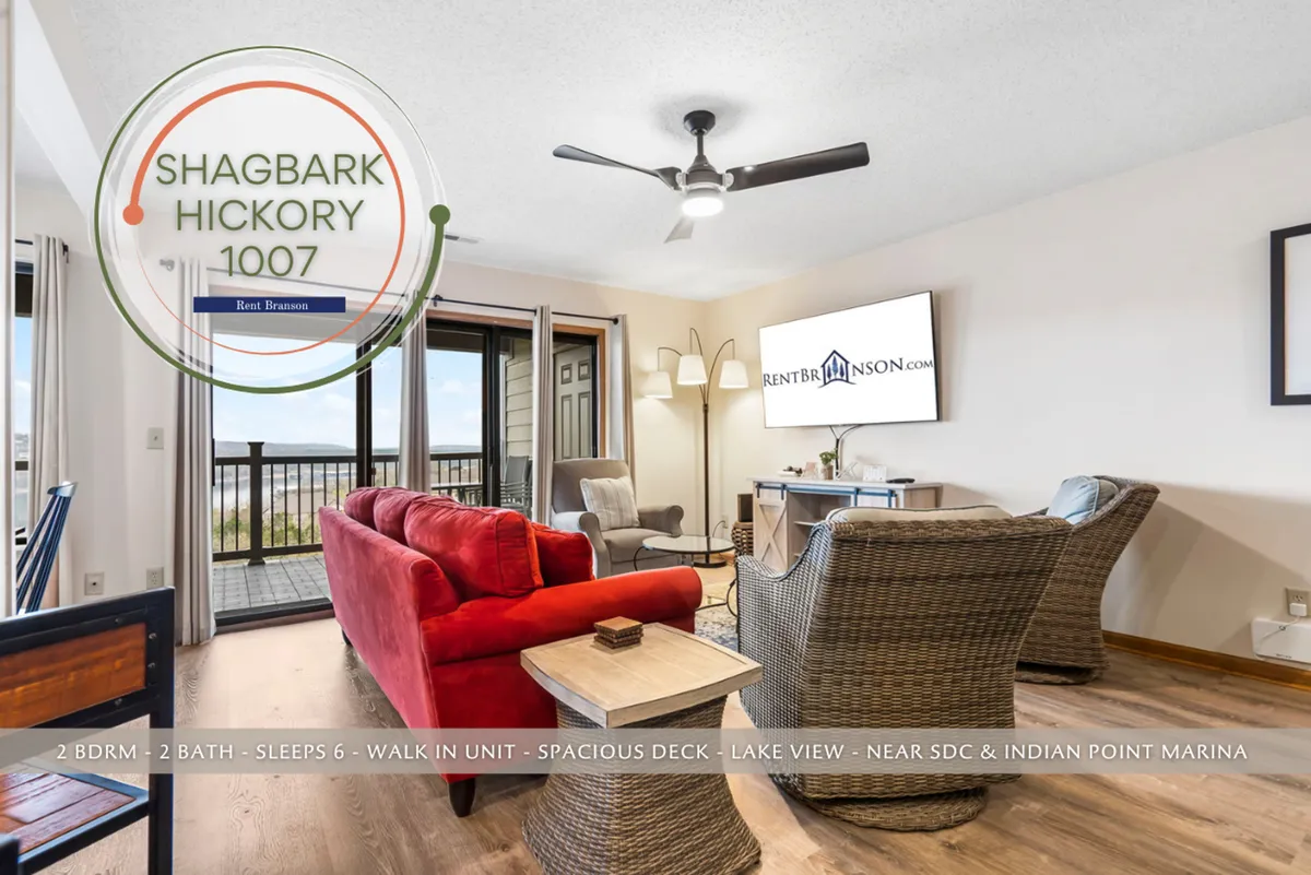 A spacious living room featuring a striking red sofa, with large windows offering a sweeping view of Table Rock Lake and a wooden deck just beyond. Warm sunlight fills the space, and the layout encourages socializing and relaxation, with board games and music accessories subtly visible.