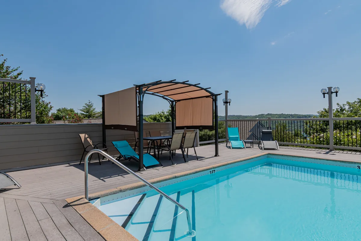 A lively outdoor pool scene with a sparkling blue swimming pool, shaded canopy seating, lounge chairs, and a backdrop of rolling landscape views. The mood is leisurely and perfect for families enjoying a summer day together.