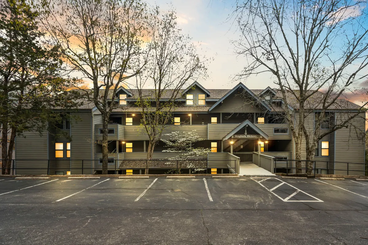 This image features the condo building's modern architecture nestled among mature trees, with warmly illuminated windows and people enjoying the balcony. The inviting exterior and visible balconies suggest spaces where families and groups can relax together, while the evening ambiance communicates tranquility and togetherness.