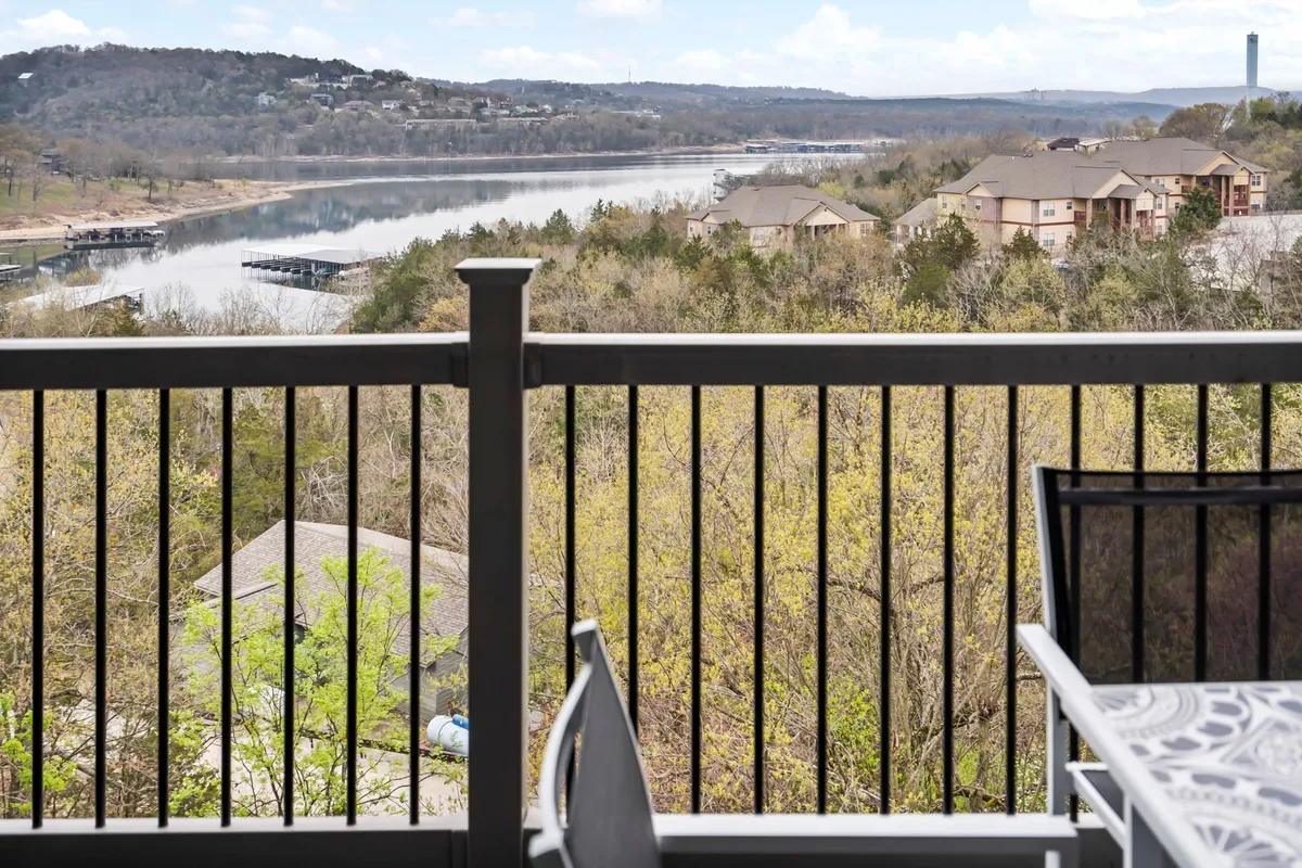 A peaceful lakeside view from a balcony or deck, with gently rippling water, lakeside homes, and lush trees. The balcony railing hints at the private relaxation space, perfect for morning coffee or unwinding in the evening while watching the sunset.