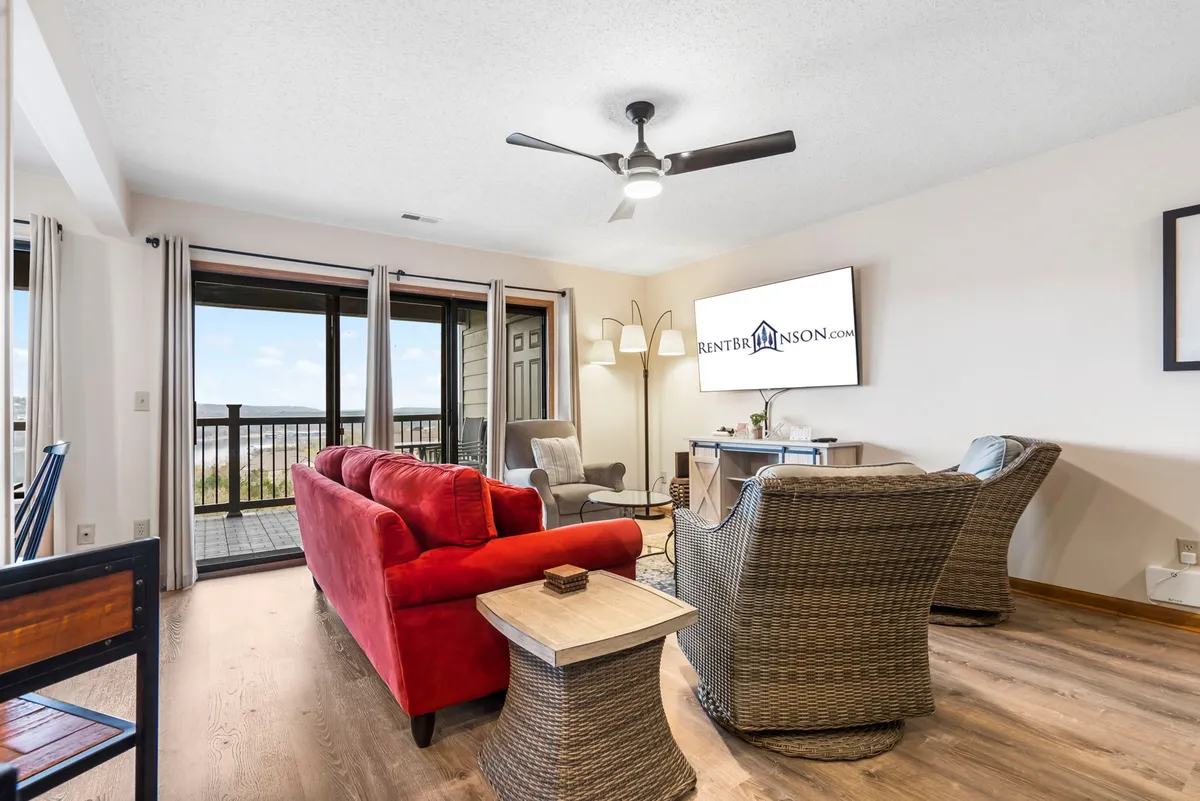 A cheerful, welcoming living space with a red sofa, wicker chairs, and large windows framing the lake. The arrangement is perfect for gatherings, with ample seating and a TV for entertainment. The sun-lit room radiates warmth and offers direct visual access to the deck and water beyond.