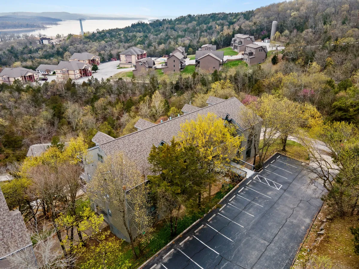 A well-maintained parking area nestled among multi-level resort buildings and shaded by green trees, with a distant view of the lake. The exterior setting highlights accessibility, ample parking, and the surrounding natural beauty. The scene feels peaceful and convenient for guests traveling with boats or trailers.