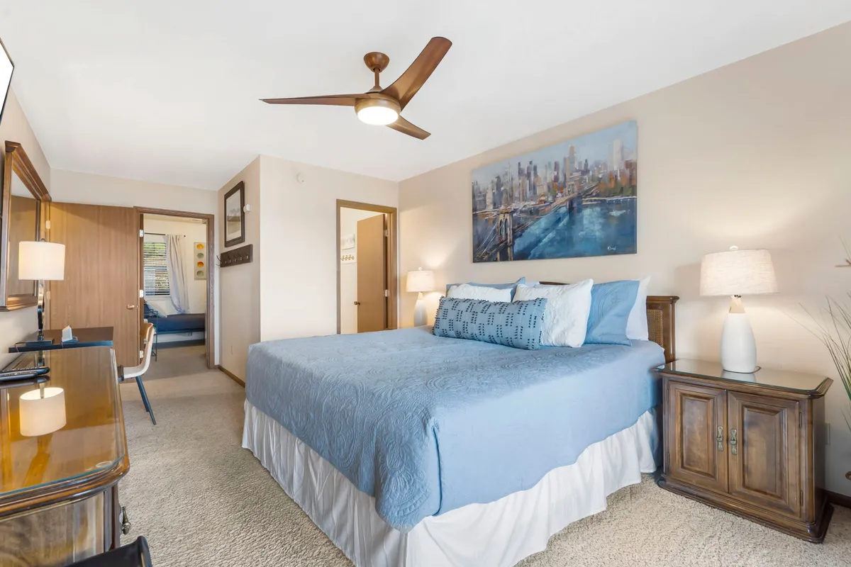 A serene and comfortable master bedroom featuring a large bed with light blue bedding, cityscape artwork above the headboard, matching wooden furniture, and a modern ceiling fan. Natural light filters in, creating a peaceful ambiance ideal for restful nights.