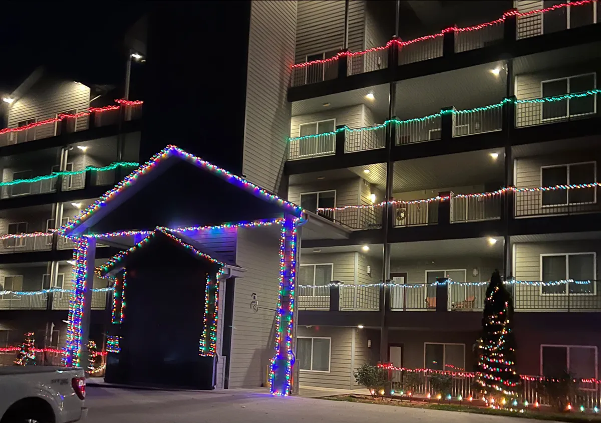 The festive exterior is adorned with colorful Christmas lights, a decorated entrance, illuminated balconies, and a Christmas tree, conveying a lively and welcoming holiday atmosphere. The setup suggests a vibrant community where guests can participate in seasonal events and celebrations.